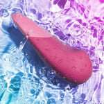 Mørk rosa Womanizer VIBE klitorisstimulator i vann med lilla og blå refleksjoner.