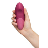 En hånd holder en mørk rosa Womanizer VIBE klitorisstimulator i silikon med ergonomisk design.