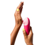 Mørke hender holder en kompakt, rosa Shine X ROMP lufttrykksvibrator med lyserosa tupp.