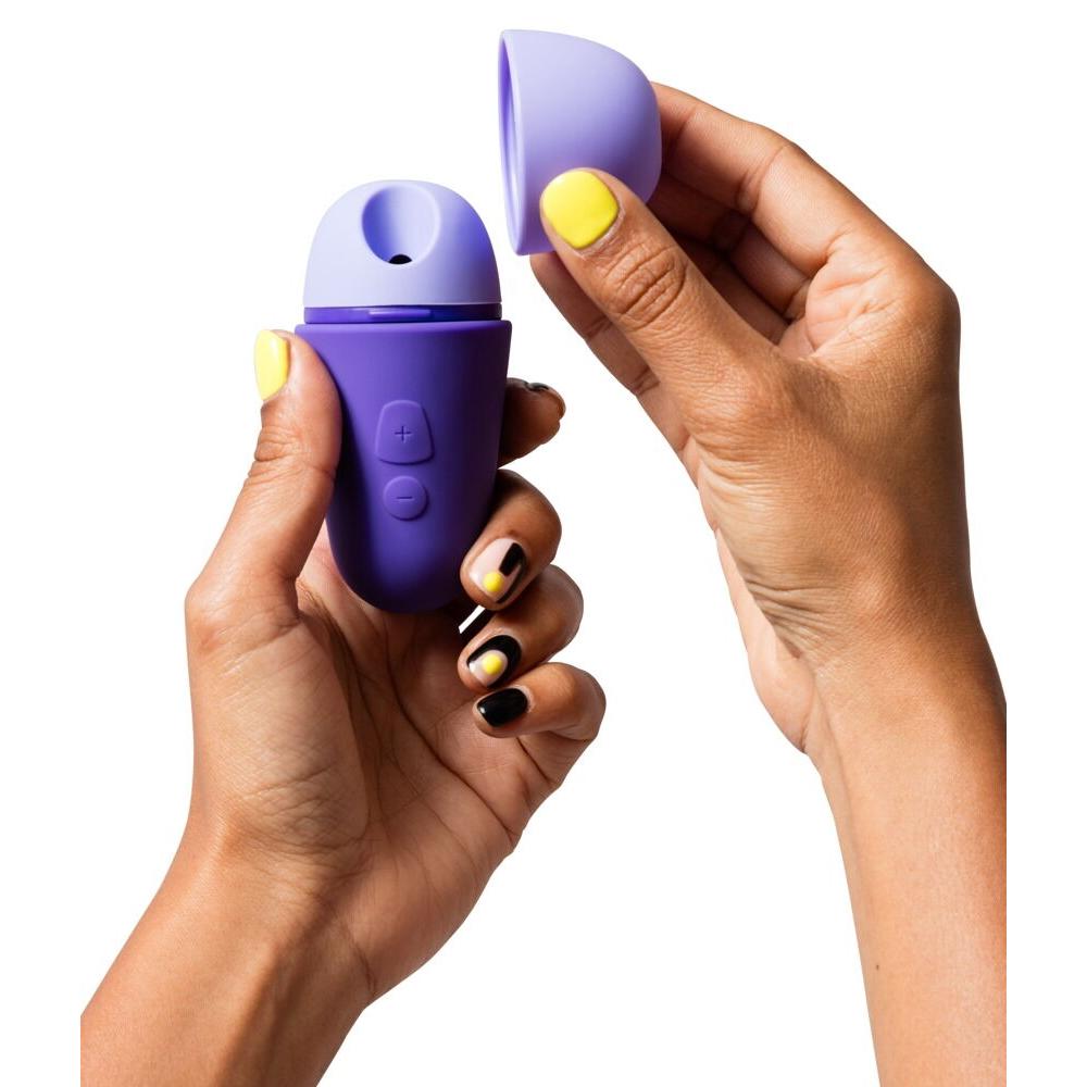Hender holder en kompakt, lilla ROMP Free X sugevibrator med åpen hette, viser sugeåpningen og kontrollknapper.