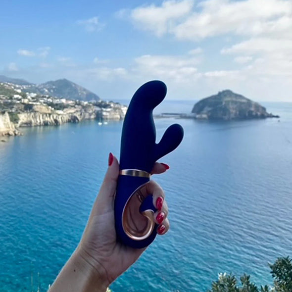 G-Vibe Gentley Rabbitvibrator i karibisk blå, med gullfarget aksent, holdt mot en havutsikt.
