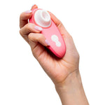 Hånd holder en korallrosa Womanizer Liberty 2 sugevibrator med hvitt stimuleringshode og kontrollknapper.