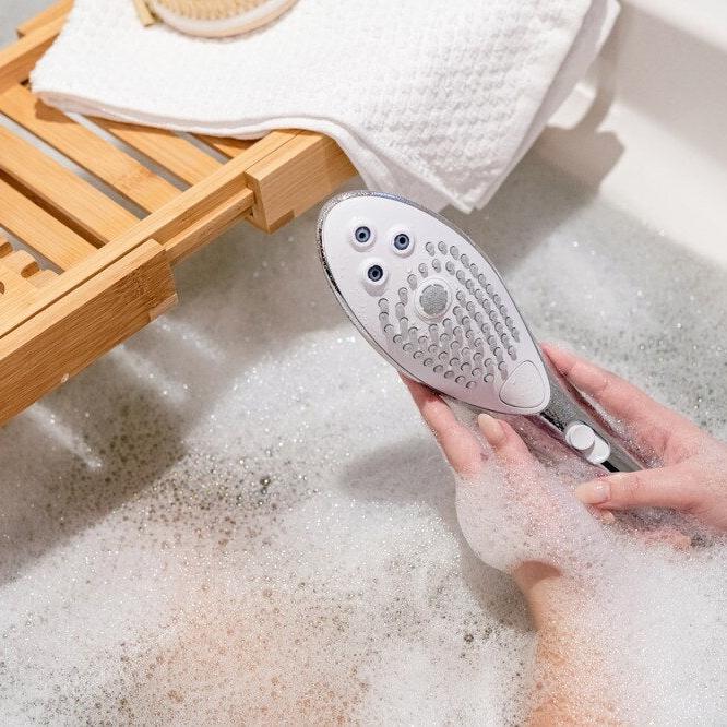Womanizer Wave dusjhode i sølv, holdt i en hånd over boblebadet, med tre stråleinnstillinger.