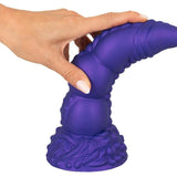 Hånd holder en lilla, teksturert Demon of Desire fantasidildo med sugekoppbase.