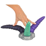 En hånd holder en Triple Tentacle fantasidildo med tre fleksible armer i lilla, grønt og mørkegrått silikon.