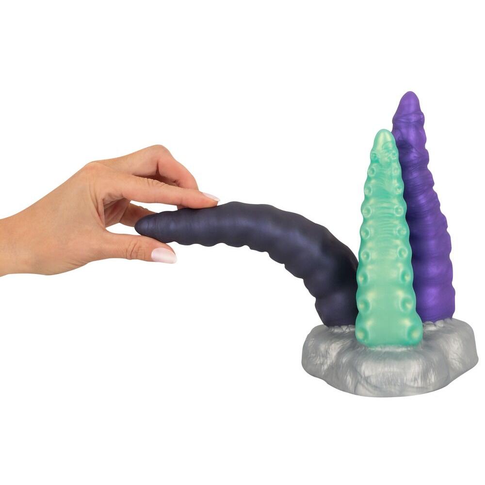 Hånd holder en mørk lilla silikon tentakeldildo med to andre i grønt og lilla på en grå base.