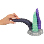 Hånd holder en mørk lilla silikon tentakeldildo med to andre i grønt og lilla på en grå base.