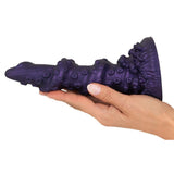En hånd holder en lilla, tentakelinspirert fantasidildo med metallisk glans og strukturert skaft.