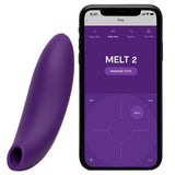 Lilla We-Vibe Melt 2 lufttrykksvibrator i silikon ved siden av en smarttelefon som viser app-grensesnittet.