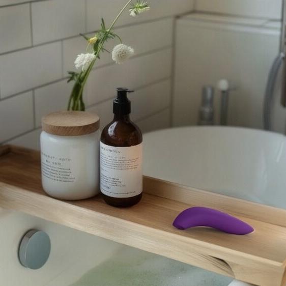 Lilla We-Vibe Melt 2 lufttrykksvibrator på et badekarbrett med baderomsprodukter og blomster.