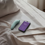 We-Vibe Melt 2 lufttrykksvibrator i dus grønn silikon, ved siden av en smarttelefon med appen.